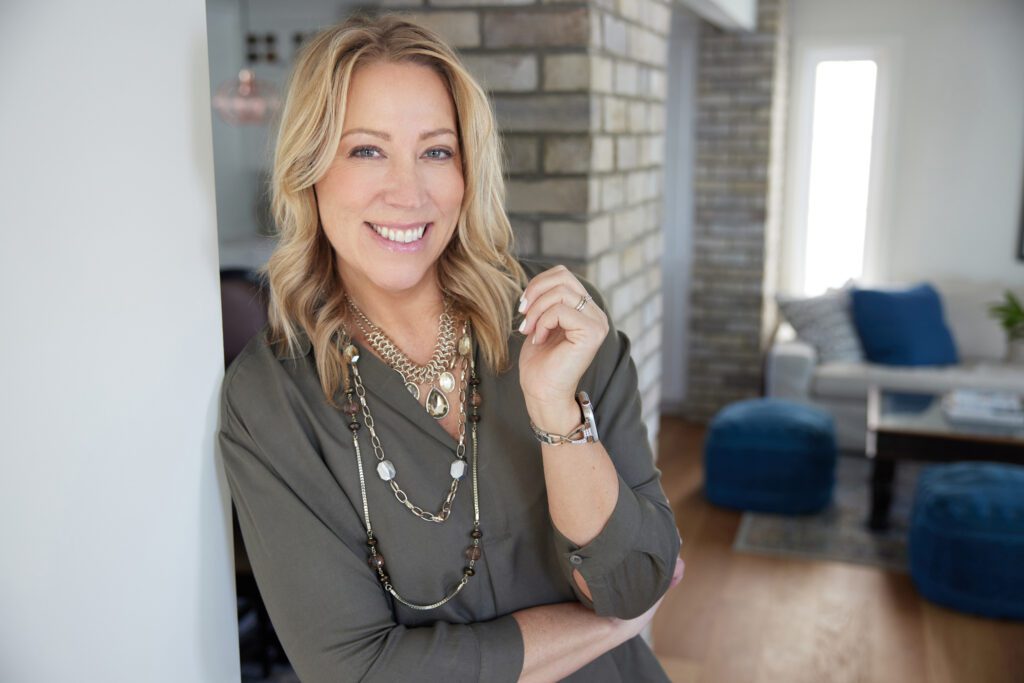 Lesley Hauville, professional Calgary real estate agent, leaning against a brick wall with a stylish modern living room backdrop, demonstrating her expertise in contemporary Calgary homes.